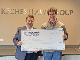 Keches Law Group partner Brian Sullivan presents an oversized check to 2021 scholarship recipient, Ronan Folan, a young man. The two stand in front of a Keches Law Group sign in an office setting.