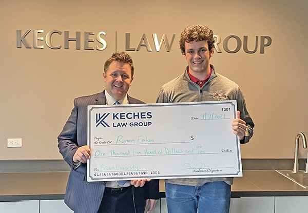 Keches Law Group partner Brian Sullivan presents an oversized check to 2021 scholarship recipient, Ronan Folan, a young man. The two stand in front of a Keches Law Group sign in an office setting.