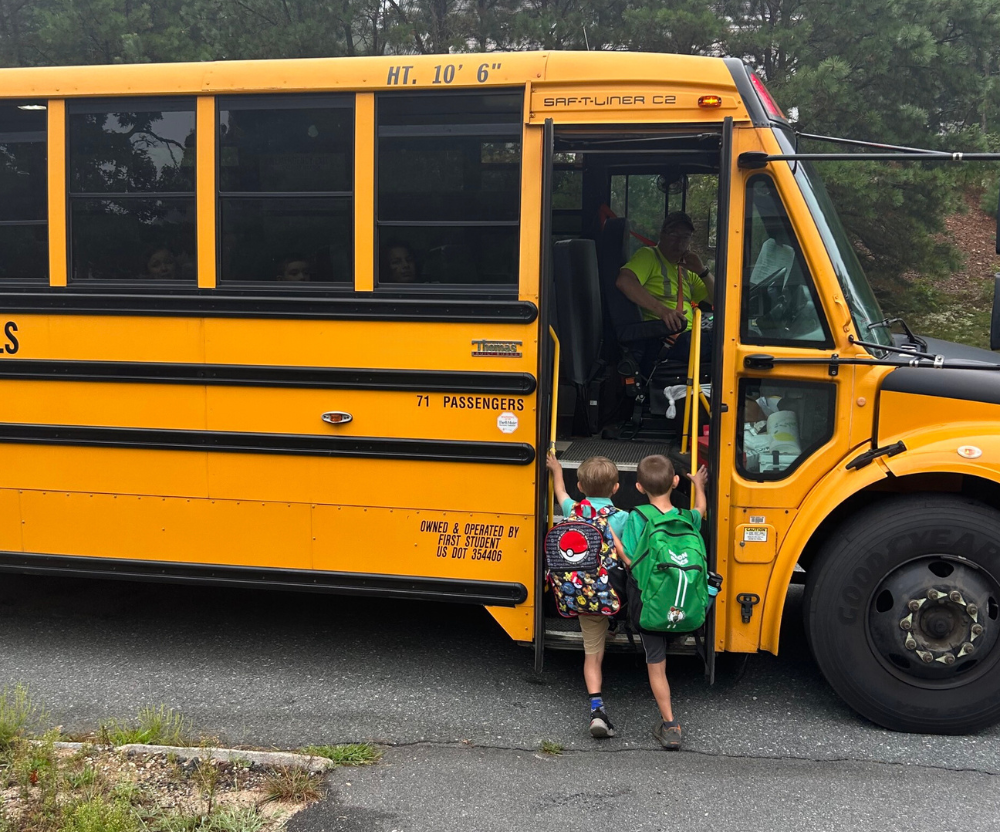 Two young boys get onto the school bus. The one on the left has what appears to be a Pokemon backpack. The one on the right has what appears to be a Boston Celtics themed backpack.