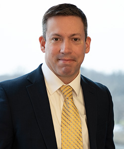 Attorney Michael Glennon, of Keches Law Group, is a Caucasian-appearing man with dark hair. He is wearing a white shirt, gold tie, and black suit jacket and is posing in front of a window.