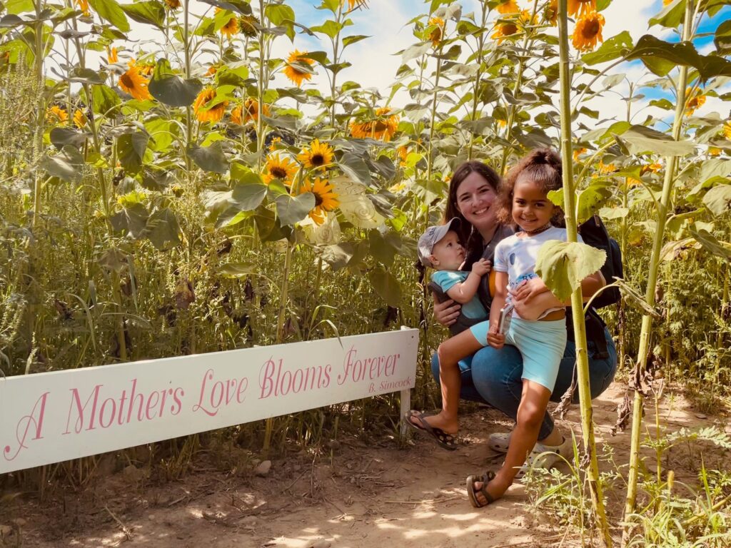 One of the working moms, a brown-haired woman named Jessica Lema, wearing jeans crouches on a path running through a field of sunflowers holding onto her two children, 1 and 1/2 year old boy appearing to be Caucasian, named John and a 4 year old girl with tan skin named Gracelynn. To her left stands a long white sign that says 'A Mother's Love Blooms Forever'.