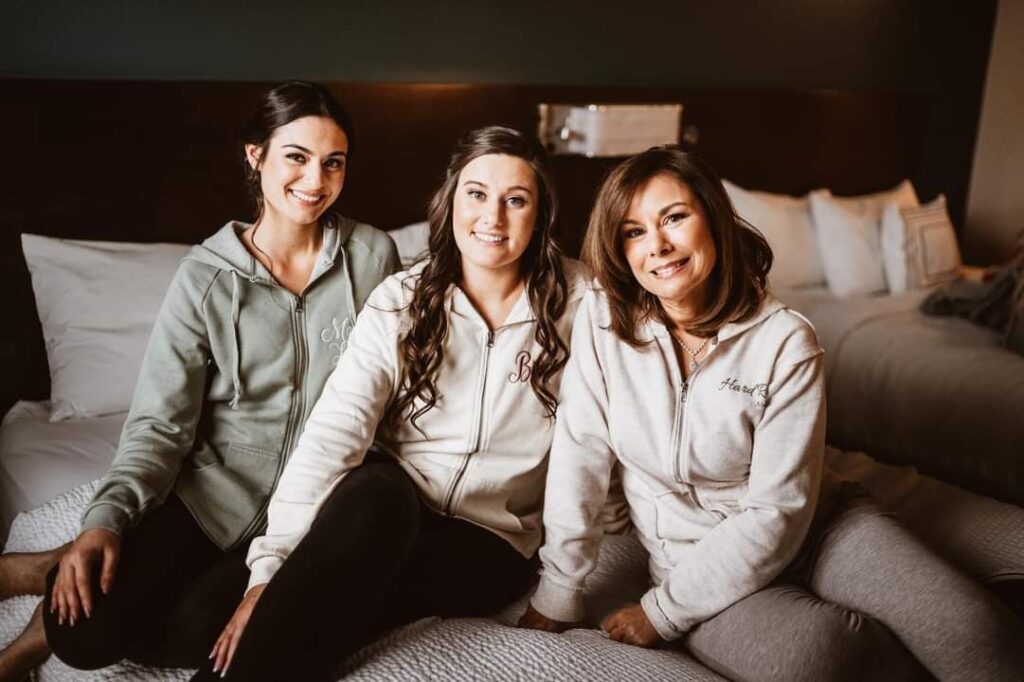 Valeri Drummond a working mom at Keches Law Group sits on a hotel room bed with her two adult children Jensen, 24, and Jessica, 28. All are wearing sweatpants and sweatshirts, have brown hair and appear to be Caucasian.