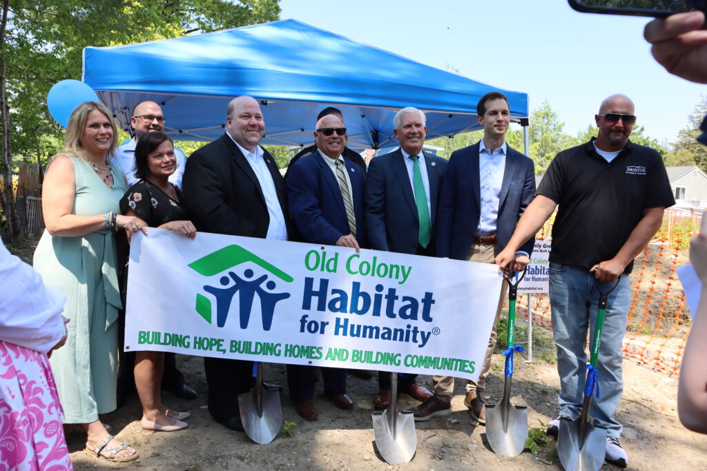 A line of representatives from Habitat for Humanity, project sponsors and politicians stand at the groundbreaking holding a sign that read "Old Colony Habitat for Humanity."