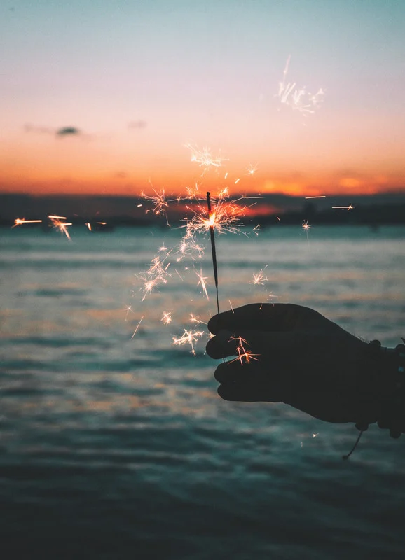 A hand holds a sparkler in front of a sunset over a body of water.