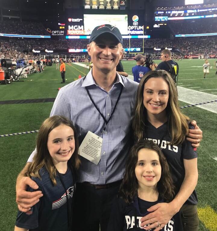 Man in Patriots hat stands with three daughters on the field at Gillette Stadium.