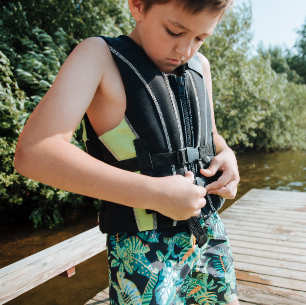 A young boy buttons up a black and green life jacket while standing on the dock in what looks like a fresh water body of water like a pond or river.