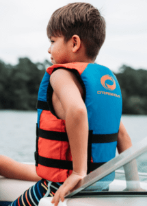 A young boy sits in a boat wearing an orange and blue life jacket.