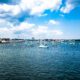A variety of sailboats anchored in the Boston Harbor in mid-June. The weather is partly cloudy with bright blue skies and a few fluffy clouds. The water is relatively calm.