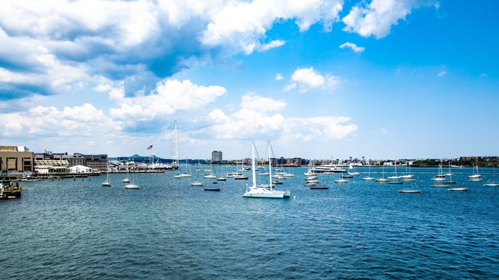 A variety of sailboats anchored in the Boston Harbor in mid-June. The weather is partly cloudy with bright blue skies and a few fluffy clouds. The water is relatively calm.