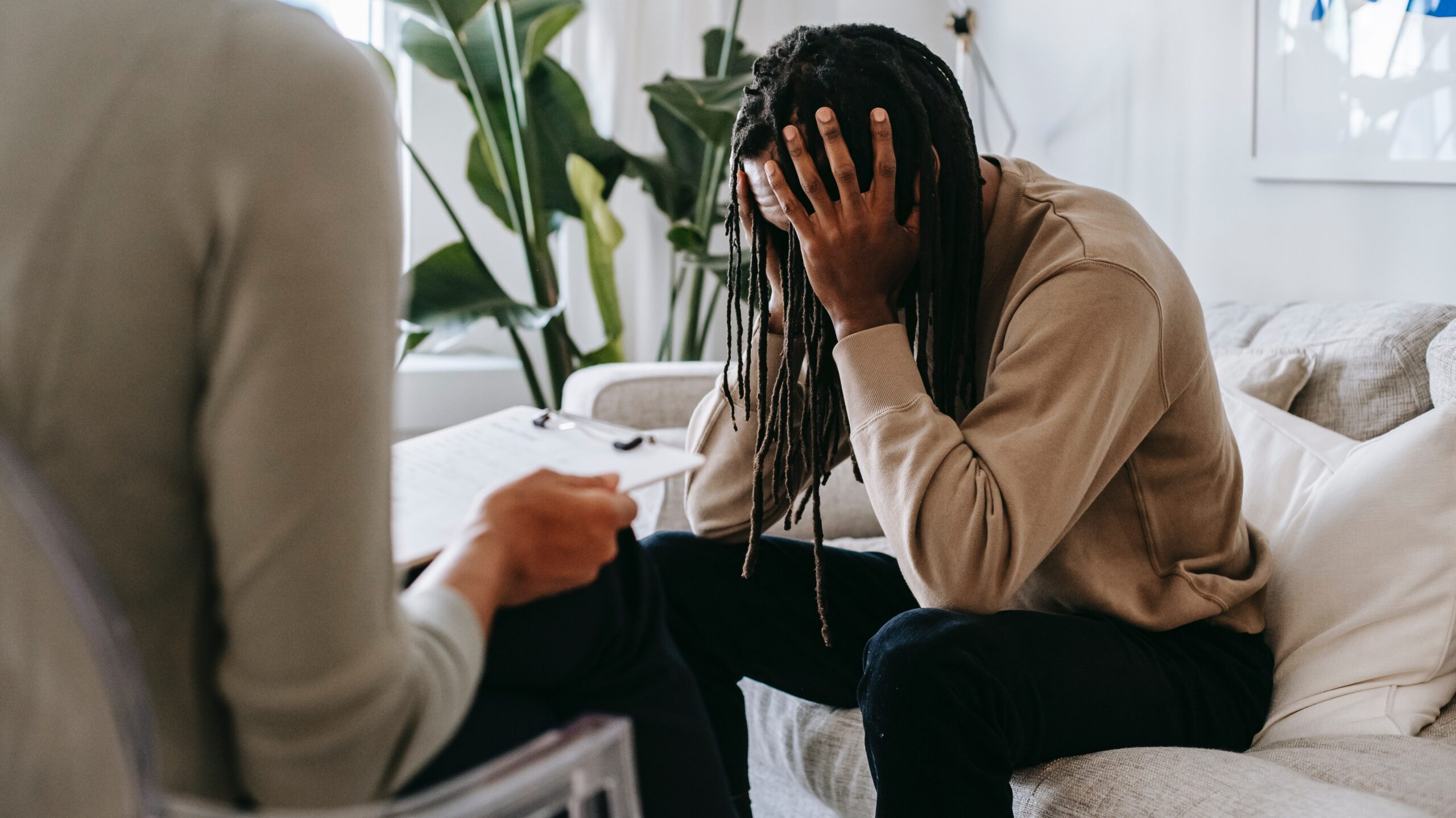 A black man with long locks wearing a tan long-sleeve shirt and black pants is sitting on a white couch holding his head in his hands while talking to a therapist.